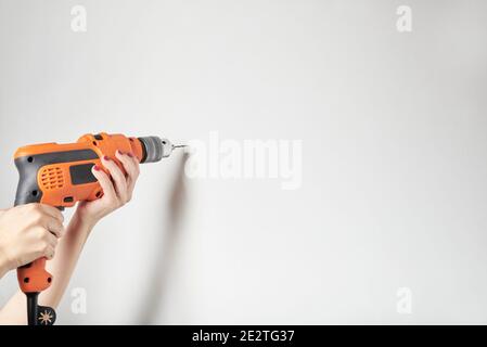 Femme aux mains manucures qui fore un mur blanc avec une perceuse électrique orange. Image détaillée avec espace de copie. Banque D'Images