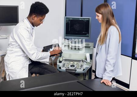 Jeune homme médecin africain dans la clinique moderne, tenant un PC tablette et montrant à sa femme caucasienne patient échographie scan des reins et interne Banque D'Images