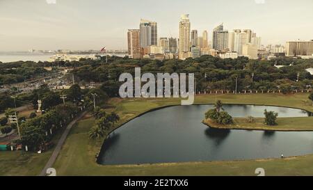 Parc vert sur la rive de l'étang en arrière-plan de soleil paysage urbain. Hauts gratte-ciels modernes sur des arbres verdoyants. Philippines métropole ville de Manille à la baie de mer. Architecture moderne et plantes tropiques Banque D'Images