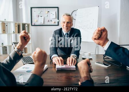 Homme d'affaires souriant tenant de l'argent près de partenaires montrant un geste oui sur premier plan flou Banque D'Images