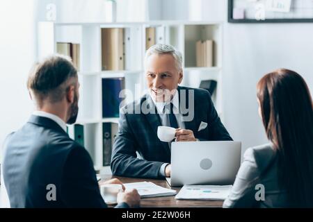 Investisseur souriant avec une tasse de café regardant les gens d'affaires avant-plan flou près des papiers et de l'ordinateur portable Banque D'Images