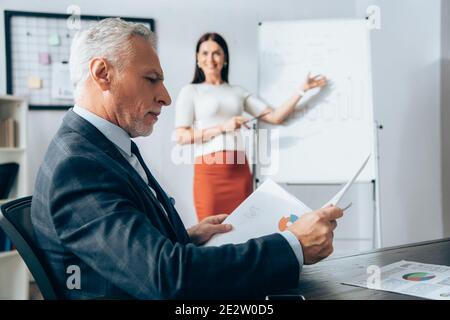 Investisseur mature regardant des documents près d'une femme d'affaires debout à côté du tableau sur un arrière-plan flou au bureau Banque D'Images