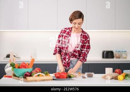 Jolie femme de ménage coupant des carottes fraîches pour le dîner de cuisine pour sa famille portant une chemise à carreaux. Cuisiner avec passion jeune femme avec les cheveux courts debout Banque D'Images