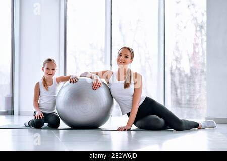 Belle jeune mère s'assoit avec sa fille sur le sol, appuyez sur le ballon de fitness à la salle de gym. Banque D'Images