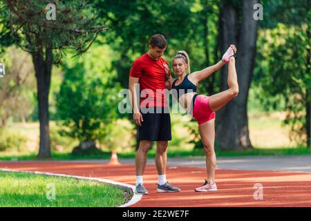 Jeune athlète masculin soutenant sa jeune femme partenaire pendant qu'elle étire les jambes après avoir courir sur une piste de course. Banque D'Images