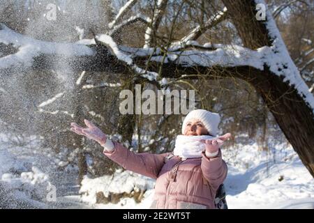 Femme vêtue d'hiver lors d'une promenade dans le parc. Jette une poignée de neige. Une rivière coule à proximité. Il y a beaucoup de neige autour. Banque D'Images