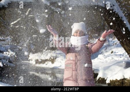 Femme vêtue d'hiver lors d'une promenade dans le parc. Jette une poignée de neige. Une rivière coule à proximité. Il y a beaucoup de neige autour. Banque D'Images