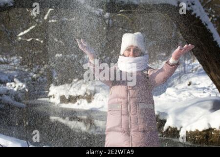 Femme vêtue d'hiver lors d'une promenade dans le parc. Jette une poignée de neige. Une rivière coule à proximité. Il y a beaucoup de neige autour. Banque D'Images