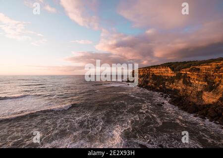 Heure d'or sur les falaises de Sao Lourenco, Ericeira, Portugal avant le coucher du soleil. Nuages dans un ciel bleu, falaises au-dessus de la mer, vagues. Banque D'Images