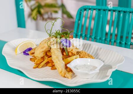 Poisson et frites avec sauce et citron, décoré de fleurs de pois papillons. Servi sur une plaque en céramique blanche. Banque D'Images