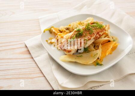 Filet de poisson de morue sur un légume de fenouil sauté avec des tranches d'orange et du poireau servi sur une assiette blanche et une serviette, table en bois clair avec espace pour les copies, sélection Banque D'Images