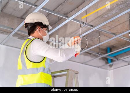 Électricien portant un masque installation pose des câbles électriques au plafond avec des pinces à l'intérieur de la maison en construction. Banque D'Images