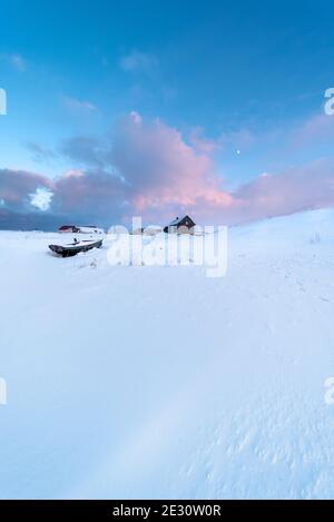 Bateau en bois et maison en bois sur un terrain couvert de neige dans un paysage arctique au coucher du soleil avec la lune dans le ciel Banque D'Images