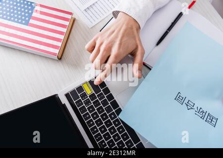 vue partielle de l'interprète en appuyant sur la touche de traduction de l'ordinateur portable à proximité papier avec hiéroglyphes et dictionnaire anglais américain Banque D'Images