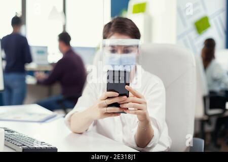 Femme d'affaires discutant et discutant sur un smartphone portant un masque facial contre le covid19 pendant une pandémie mondiale. Collègues multiethniques travaillant dans le respect de la distance sociale dans la société financière. Banque D'Images