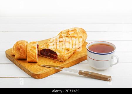 Strudel de cerisier maison parsemé de noix écrasées sur une planche à découper près d'un couteau de cuisine et d'une tasse de thé sur une table en bois blanc. Banque D'Images