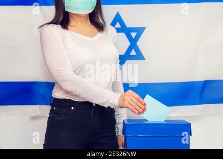 Femme israélienne avec masque médical à visage protégé sur fond de drapeau israélien mettant le vote dans une boîte de vote le jour du scrutin. Banque D'Images