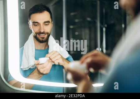 Gros plan de beau jeune homme avec une serviette dans son cou debout devant le miroir et appliquant de la crème hydratante sur son visage. Banque D'Images