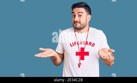 Jeune homme beau portant l'uniforme de maître-nageur expression sans indice et confuse avec les bras et les mains soulevées. Doute concept. Banque D'Images