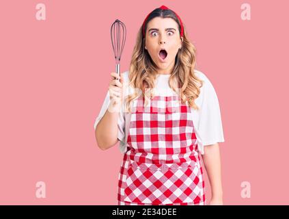 Jeune femme caucasienne portant un tablier tenant le fouet effrayé et stupéfié avec la bouche ouverte pour la surprise, incrédulité visage Banque D'Images