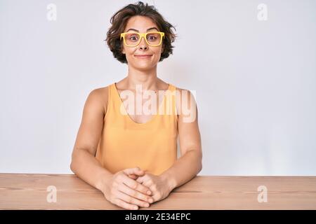 Jeune femme hispanique portant des vêtements et des lunettes décontractés assis sur la table puffant les joues avec le visage drôle. Bouche gonflée avec de l'air, expression folle Banque D'Images