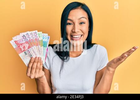 Belle femme hispanique détenant des billets de banque en dollars de hong kong célébrant la réussite sourire heureux et expression du gagnant avec main levée Banque D'Images