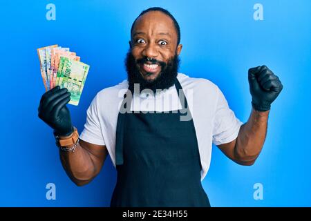 Un jeune homme afro-américain portant un tablier professionnel tenant des billets de rand en afrique du Sud criant fier, célébrant la victoire et le succès très excite Banque D'Images