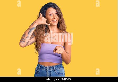 Jeune femme hispanique avec tatouage portant des vêtements décontractés souriant en parlant sur le geste de téléphone et vous pointant vers vous. Appelez-moi. Banque D'Images