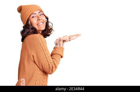 Jeune belle femme de course mixte portant un chandail en laine et un chapeau d'hiver pointant de côté avec les mains ouvertes palmiers montrant l'espace de copie, présentant la publicité Banque D'Images