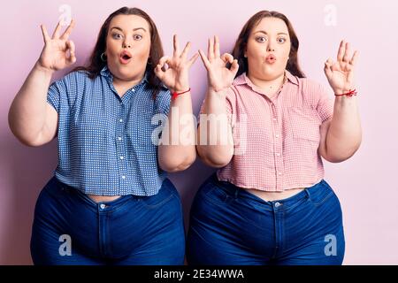 Jeunes jumeaux de taille plus portant des vêtements décontractés ayant l'air surpris et choqué faisant ok symbole d'approbation avec les doigts. Expression folle Banque D'Images