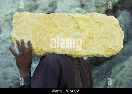 Le mineur porte le bloc de soufre dans les mines de soufre du cratère du volcan actif de Kawah Ijen à Java-est, en Indonésie. Chaque matin, deux centaines de mineurs descendent jusqu'au fond du cratère pour la prochaine charge de soufre. Ils remplissent leurs paniers avec du soufre manuellement et portent ensuite cette lourde charge sur les pieds. Le travail extrêmement dur dans les nuages de dioxyde de soufre toxique provoque une toux dure et des yeux qui tournent presque immédiatement. Cependant, les mineurs travaillent sans protection. Peu d'entre eux ont des masques à gaz. Chaque mineur gagne environ 10 dollars américains par jour. Banque D'Images