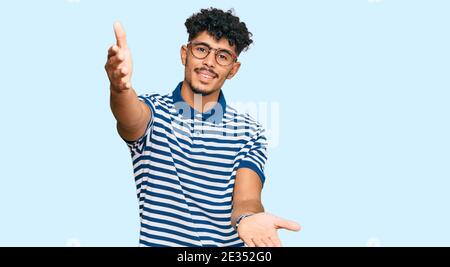 Jeune homme arabe portant des vêtements et des lunettes décontractés regardant la caméra souriant avec les bras ouverts pour le câlin. Expression gaie embrassant le bonheur. Banque D'Images