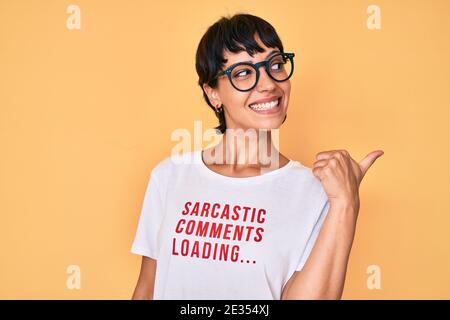 Belle femme brunettte portant des commentaires sarcastique chargement du t-shirt sourire avec le visage heureux regardant et pointant vers le côté avec le pouce vers le haut. Banque D'Images