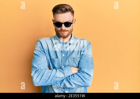 Jeune homme à tête rouge portant des lunettes de soleil stylées sceptique et nerveux, désapprouvant l'expression sur le visage avec les bras croisés. Négatif personne. Banque D'Images