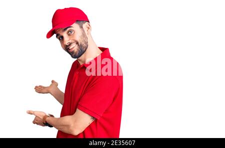 Jeune homme beau avec barbe portant un uniforme de livraison invitant à entrez dans le sourire naturel avec la main ouverte Banque D'Images