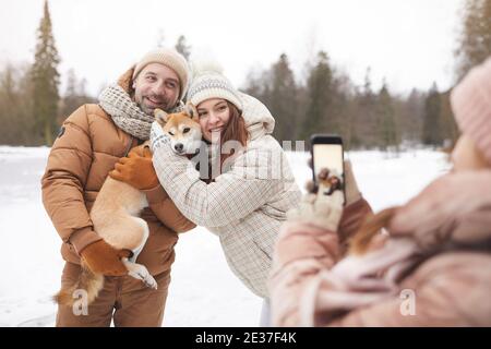 Adolescente prenant des photos de parents tout en appréciant marcher dehors ensemble dans la forêt d'hiver, se concentrer sur le couple heureux adulte tenant le chien Banque D'Images