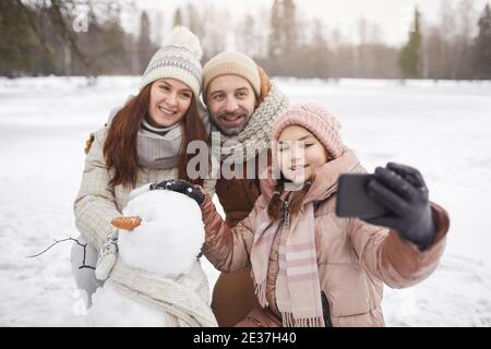 Portrait d'une famille heureuse prenant des photos de selfie à l'extérieur tout en construisant un bonhomme de neige ensemble et en profitant de vacances d'hiver, espace de copie Banque D'Images