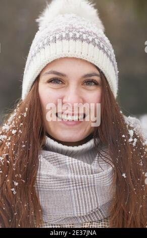Portrait vertical en gros plan de la jeune femme souriante en hiver porter un chapeau en tricot tout en regardant l'appareil photo recouvert de neige Banque D'Images