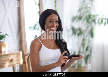 Cosmétiques et beaux cheveux longs sains. Souriant, une jeune femme afro-américaine prend soin de ses cheveux Banque D'Images