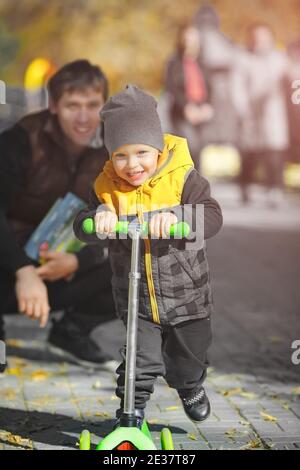 Un petit garçon heureux sur un scooter marche dans le parc avec son père un jour d'automne. Banque D'Images