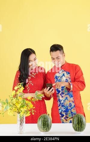 Jeune couple asiatique en robes traditionnelles debout à la table avec des pastèques fraîches et l'envoi de messages texte aux membres de la famille sur Nouvel an chinois Banque D'Images