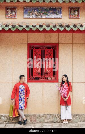 Petit ami et petite amie dans les robes vietnamiennes traditionnelles debout à l'extérieur avec fleurs et sacs de shopping et regardant l'un l'autre Banque D'Images