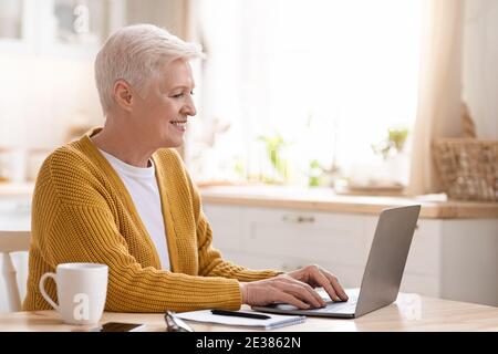 Femme aîée gaie assise dans la cuisine, à l'aide d'un ordinateur portable Banque D'Images