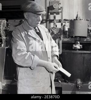 dans les années 1950, historique, un homme âgé qui porte un ouvrier à bouton avant ou un manteau de coton d'entrepôt et un chapeau de tissu, examine de près une carte qu'il tient. Peut-être un tableau de couleurs pour le mélange de peinture. Son manteau est un uniforme de travail traditionnel, connu sous le nom de manteau d'entrepôt et étaient avant fesses et avait deux poches basses patch et une poche supérieure patch et étaient destinés aux travailleurs de la capentérale ou de l'industrie générale, en gardant les vêtements sous-jacents propres. Banque D'Images