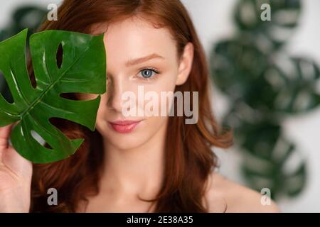 Jeune femme aux cheveux rouges posant avec une feuille tropicale sur fond blanc Banque D'Images