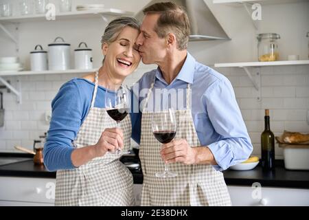 Joyeux couple plus âgé qui s'enserre, qui boit du vin dans la cuisine pour célébrer la Saint-Valentin. Banque D'Images