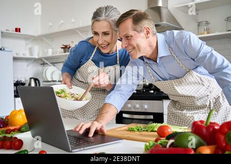 Un vieux couple heureux utilisant un ordinateur portable pour préparer une salade végétalienne à la maison dans la cuisine. Banque D'Images