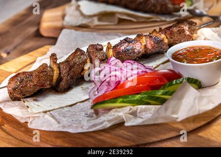 Brochette de porc sur une brochette de métal avec sauce, tranches de concombres, tomates et oignons sur un plateau en bois Banque D'Images