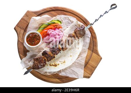 Brochette de porc sur une brochette de métal avec sauce, tranches de concombres, tomates et oignons sur un plateau en bois sur fond blanc isolé. Banque D'Images