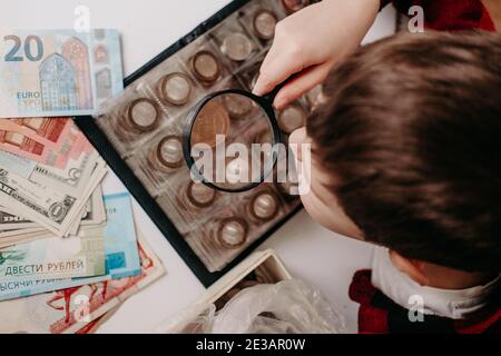 collecte de pièces. Le garçon examine une vieille pièce à travers une loupe. Dollars, euros sur la table. Argent de différentes nations. Économies dans le for Banque D'Images